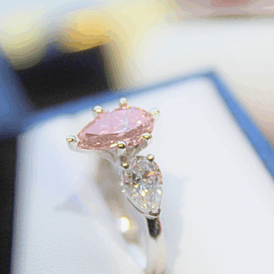 Ring with a 1.54 ct pink pear-shaped diamond center accented by two white pear-shaped diamonds, set in 14K white gold, displayed on a ring stand with a sparkly background.